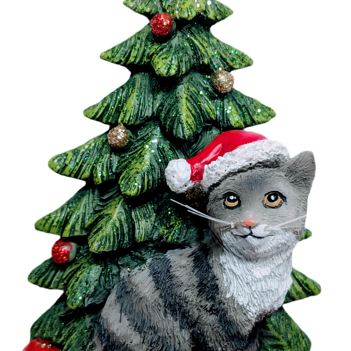 Grey Cat Sitting By Christmas Tree Wearing A Red Santa Hat Ornament